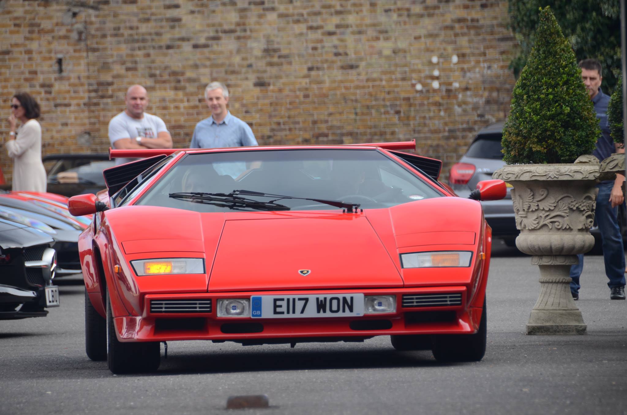 Sheesh Supercar lunch II - Sunday 21st September 2014 - Page 2 - Supercar General - PistonHeads - This image features an eye-catching red Lamborghini car on a busy street. The Lamborghini, with its sleek design and iconic logo, is attracting attention as it drives away from a group of people and other cars, which are partially visible in the background. These individuals appear to be admiring the car, creating a scene where the car seems to be the star of the show. The street where this all is taking place looks urban, and there are multiple other vehicles adding to the bustling atmosphere.