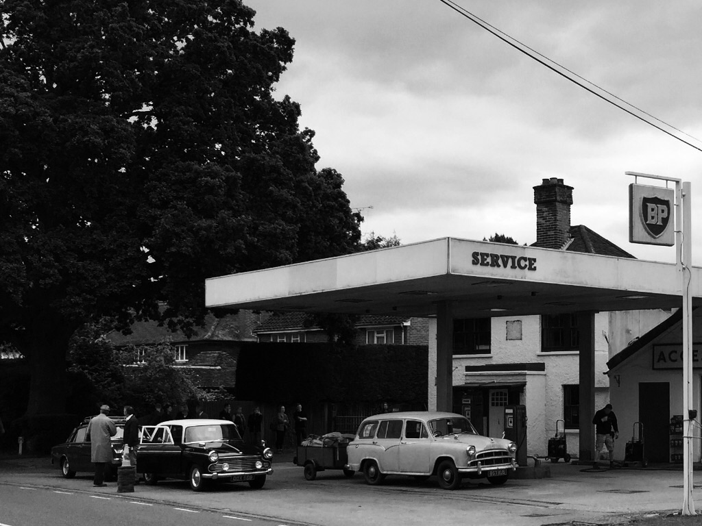 Random Photos : Part 4 - Page 204 - Photography & Video - PistonHeads - The image is a black and white photograph of a roadside scene. At the center of the frame is an old-fashioned gas station named "Service" with "BP" visible on an additional fixture attached to the station. Surrounding the gas station are several cars parked in the station's lot. The street where the cars are parked is bordered by lush green trees, adding a touch of nature to the urban setting. A prominent tree next to the gas station suggests a well-maintained green space in the vicinity.