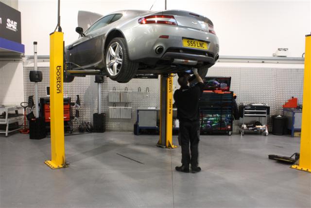 V8 Vantage tuning (SLINC) - TDI Inspection Service - Page 1 - Aston Martin - PistonHeads - The image portrays a scene in a service garage or workshop. In the forefront, there is a man standing on a lift to perform maintenance or repairs on a car. He is engrossed in his work, focused on adjusting the car. The lift is attached to a sturdy yellow post. The surroundings reveal tools and equipment, suggesting a professional automotive environment. In the background, a clock can be seen, possibly indicating the service rate or appointment time for the car.