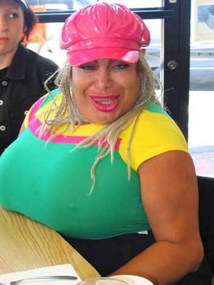 Pistonheads - The image depicts a woman sitting at a table in a restaurant. She is wearing a vibrant and unusual outfit, including a pink hat, a green, pink, and yellow top, and a leafy green wig that spills over her shoulders, swishing across her chest. Her face is exaggeratedly painted, with pink lipstick and an extra layer of foundation on her cheeks, adding a dramatic touch to the overall look. On the table in front of her, there is a plate with an unseen item on it, suggesting she might be eating or about to order something. To the left of the frame, there appears to be a door, and in the background, another person can be seen, though only the upper body and head are visible.