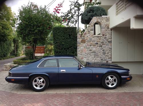 A car parked in front of a building - Pistonheads - The image displays an old-style, blue-colored coupe parked alongside a paved driveway of a house. The car is positioned against the curb, facing towards the right. The house features a stone facade with a pillar and a small rectangular window. A lush green hedge stands in the background, partially framing the car. The scene suggests a residential area with well-maintained landscaping.