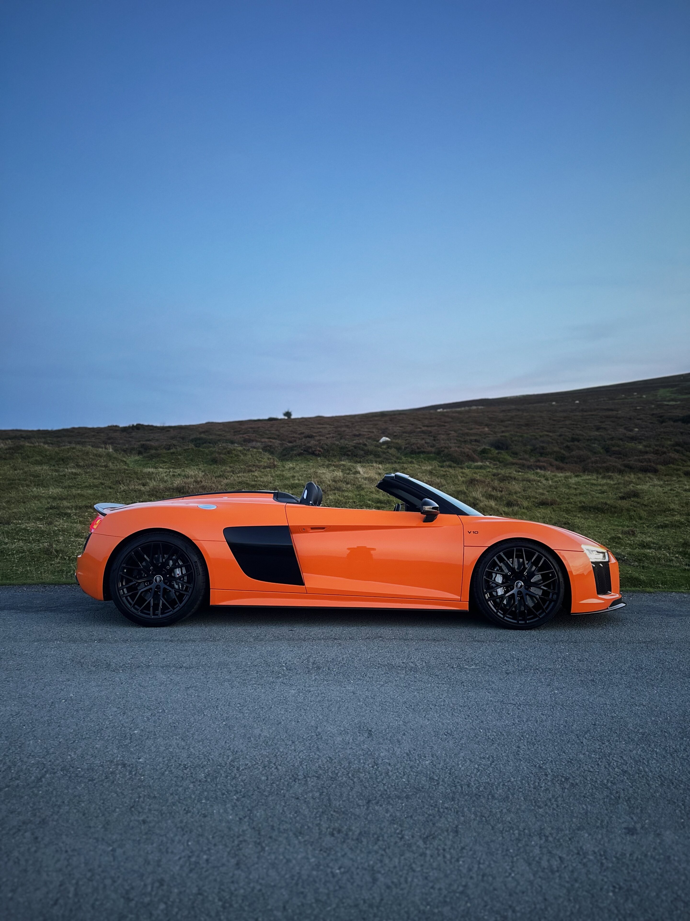 Orange Audi R8 V10 Plus Spyder  - Page 2 - Readers' Cars - PistonHeads UK - This image captures a scene on a road at dusk. A vibrant orange sports car, possibly an Audi RS or similar, is parked on the side of the road. The car's sleek design and striking color make it stand out against the natural backdrop of a hillside. There are no people visible in the image. The sky above is painted with hues of blue, suggesting that the photo was taken either early in the morning or late in the afternoon. The setting sun casts a warm glow on the scene, adding to its serene ambiance.