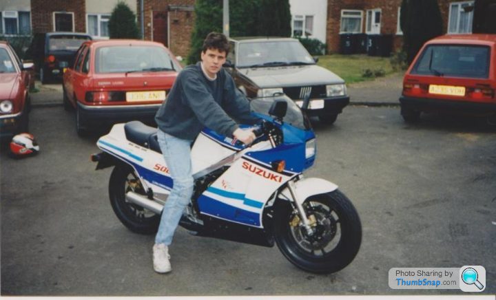 A man riding on the back of a motorcycle - Pistonheads - The image showcases a person sitting on a blue and white motorcycle. The motorcycle is white and blue with the word "SUZUKI" visible on it. The person is wearing a helmet, suggesting they may be preparing for a ride. The motorcycle appears to be parked in an area with other parked cars visible, indicating that the location might be a parking area or a driveway. The person's posture and the position of the foot on the footstand suggest they are relaxed and possibly posing for the photo.