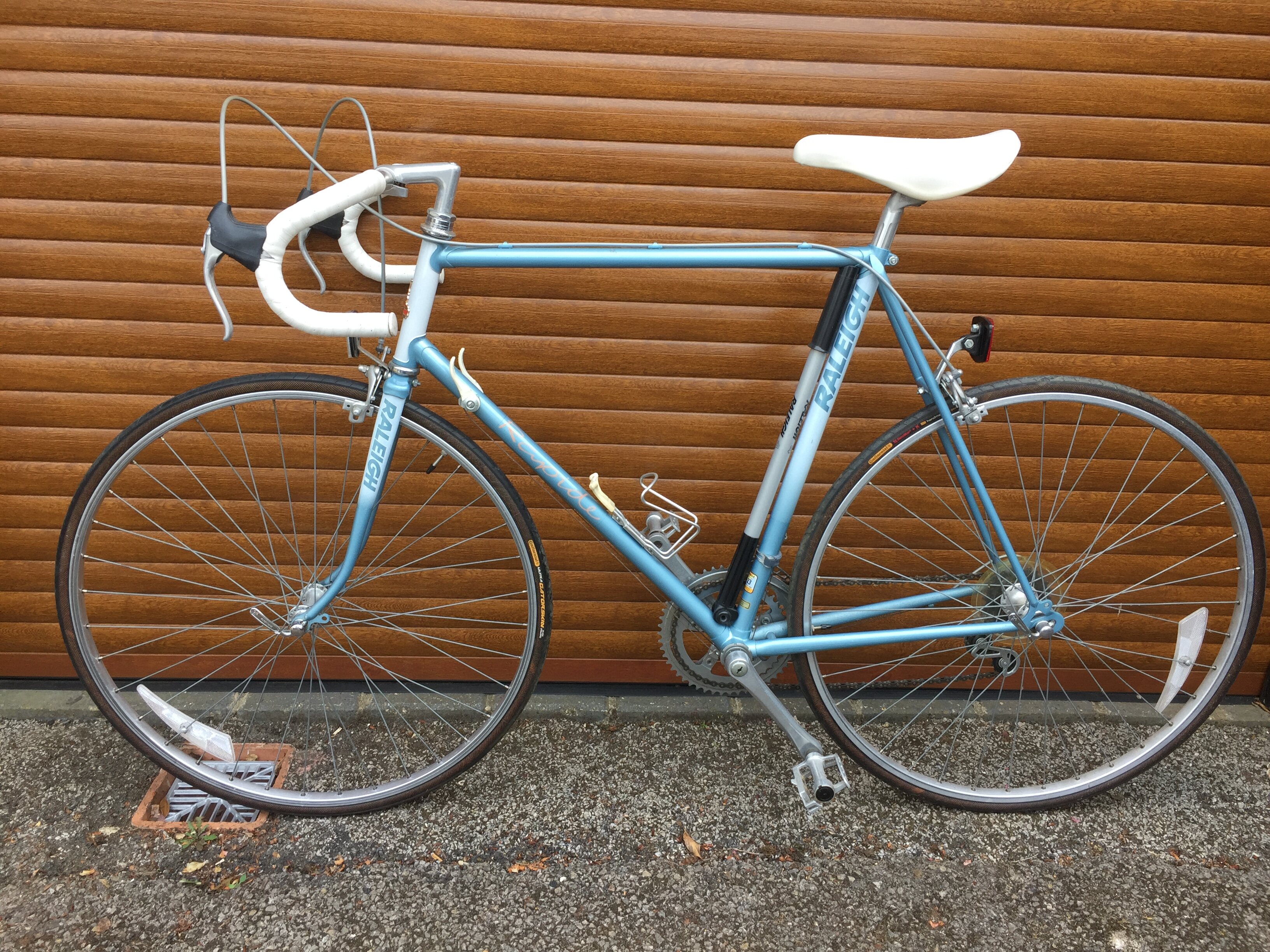 you nostalgia you lose - Page 148 - The Lounge - PistonHeads UK - The image showcases a vintage ten-speed bicycle, parked against a garage door. The bicycle is painted in a distinctive shade of blue and features a white seat for comfort. The frame of the bicycle appears sturdy and well-maintained, indicative of many hours of riding. In the background, you can see a building with a wooden exterior, providing a rustic setting that complements the old-fashioned style of the bike.