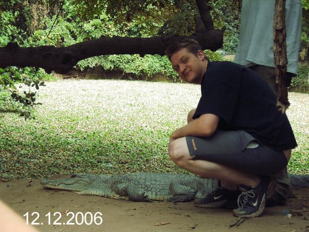 In this image, a man is sitting on the ground next to a large alligator. He is wearing a black t-shirt and gray shorts with a white logo on them. The alligator is gray with a long snout and large eyes, and it appears to be at rest within its enclosure. The scene takes place in what appears to be a green vegetated area with a tree nearby. The date stamp on the picture indicates it was taken on December 12, 2006.