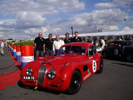 Historic Pistonheads Silverstone Xks - In the image, a group of four men are gathered around a vintage red car at a car show. The men are all wearing sunglasses and are standing behind the car. The vintage red car bears the number "5" on it. The car show appears to be taking place in an outdoor setting, possibly a parking lot or an open field, as other vintage cars are visible in the background. The setting is bathed in bright sunlight, creating a lively and festive atmosphere for the car show.