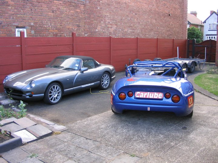 GT style racer hard top - Page 1 - Major Mods - PistonHeads - The image captures a scene in a driveway where two vintage cars are parked. One is a silver sports car with a sleek design and shiny rims, while the other is a blue convertible with a round design visible under the hood. Both cars are parked next to a red brick fence, and there's a yellow cord plugged into the sports car. The driveway shows signs of use with spots on the ground, and there's a white house faintly visible in the background. The overall atmosphere is nostalgic, evoking a sense of classic elegance.