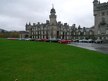 Pistonheads - This image displays a large, sprawling building with a traditional architectural style, which appears to be a castle or a similar historical structure with multiple spires and turrets. In front of the building, there is a neatly arranged line of cars, predominantly parked at an angle along the grassy ground; all cars are positioned facing the same direction. The cars are varied in color, model, and make, adding a modern contrast to the historic building in the background. The sky overhead is overcast, suggesting a cloudy or possibly rainy weather condition. The overall impression is of a large, historic site, now serving as a temporary parking lot for visitors.