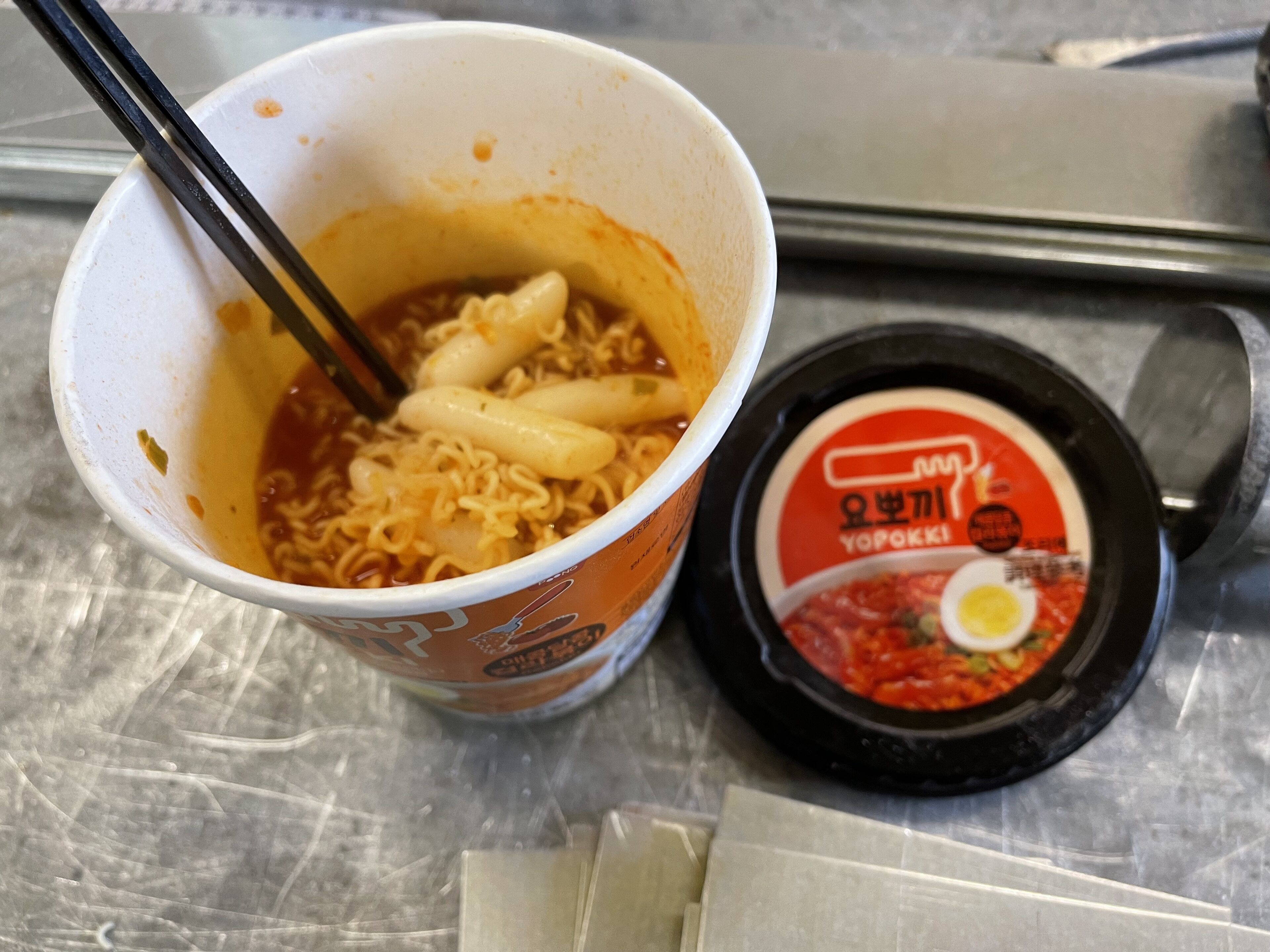 noodles in a pot - Page 25 - Food, Drink & Restaurants - PistonHeads UK - The image shows a meal set on a table. In the foreground, there's a container with noodles and broth, accompanied by a packet of condiments labeled "Taeyoung," which translates to "Red Ginseng." Adjacent to this container is a small, cylindrical package that appears to contain some sort of spice or seasoning. Behind these items, there's a disposable cup with a lid, containing what looks like instant noodles, possibly in a soup base. The meal is presented in what seems to be a takeaway setting, as suggested by the packaging and utensils present.