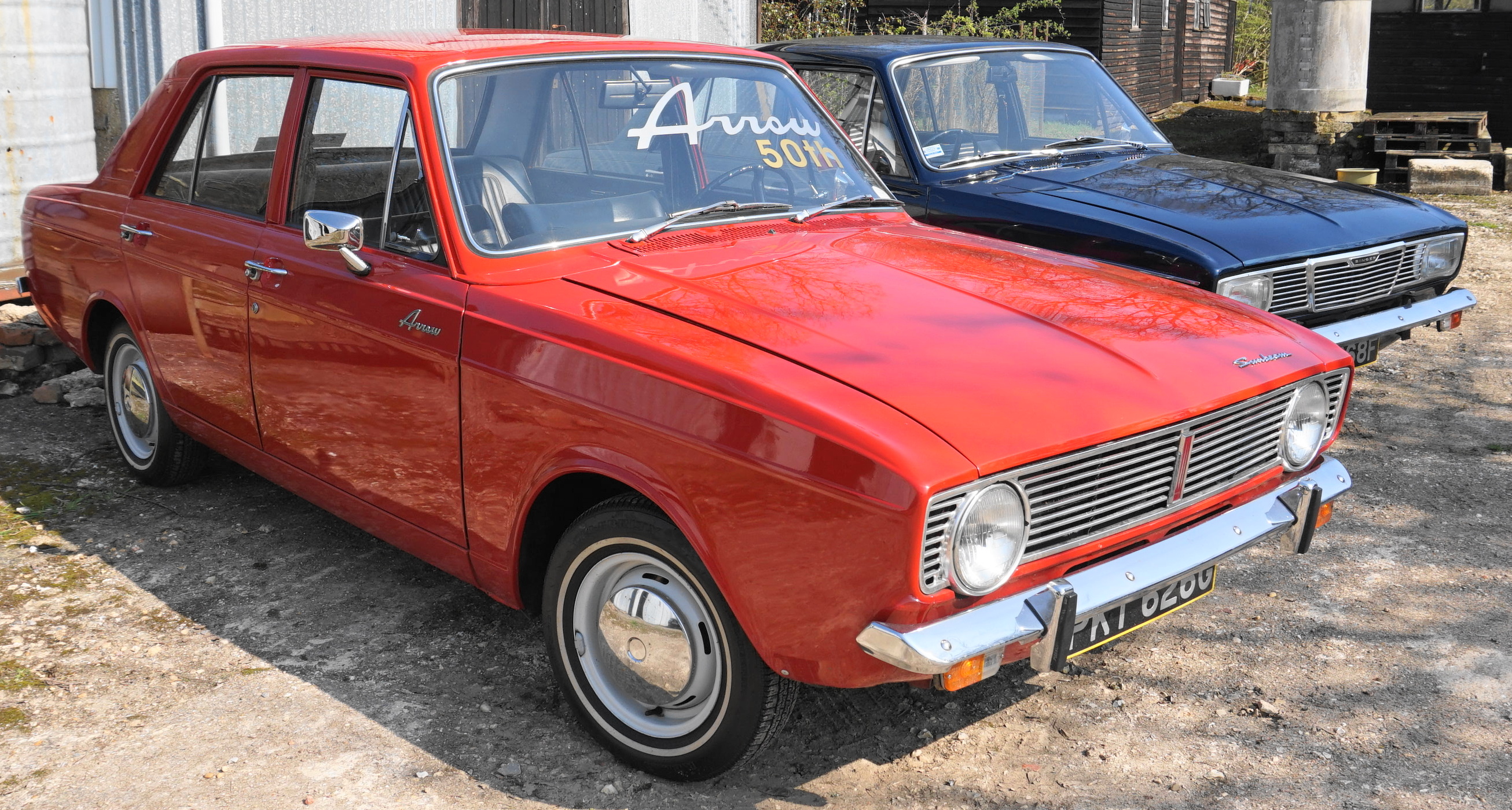 Mystery boat engine, possibly 1960s Ford Anglia ? - Page 1 - Classic Cars and Yesterday's Heroes - PistonHeads UK - The image shows two classic cars parked side by side on a gravel surface. The car on the left is an older model, likely from the 1950s or 60s, characterized by its prominent fins and chrome details. It has a red body with a cream-colored interior. The car on the right appears to be of similar vintage but may not have the same level of detail or restoration as the first car. Both cars are parked in an outdoor setting, possibly a showroom or exhibition area dedicated to classic vehicles.