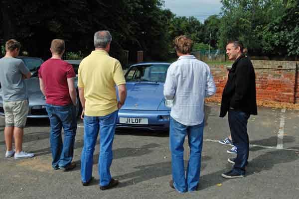 A group of men standing next to each other - Pistonheads - The image shows four men standing in a parking lot with a line of cars. One man has a distinctive hairstyle. They appear to be engaged in a conversation or observation. There is foliage in the background, indicating a reasonably sunny day. The cars look like new models.