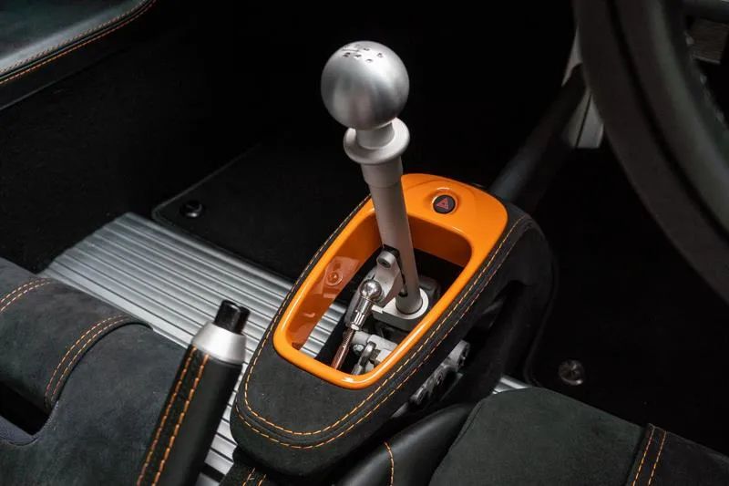 Pistonheads - The image shows the interior of a car, focusing on the gear shift area. An orange gear knob is prominently visible in the center of the photo. The car's steering wheel is visible at the top of the frame, indicating it's mounted on a column. A black and tan seatbelt spans across the image, adding to the sense of motion within the vehicle. The gear shift is situated on a racing-style console that is integrated into the center console of the car. The background suggests a well-lit interior, likely in a garage or showroom.