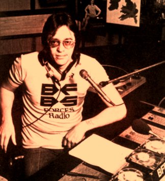 Pistonheads Albums Swansong - This is a black and white photograph featuring a man seated at a radio broadcasting desk. The man is wearing headphones and glasses, and he is smiling at the viewer. He is dressed in a white t-shirt with a graphic and text design on it. The text on his shirt reads "B5G" and "FORCES RADIO". His hands are on the desk in front of him, and there are several controls and meters in front of him, indicative of a radio broadcasting station.