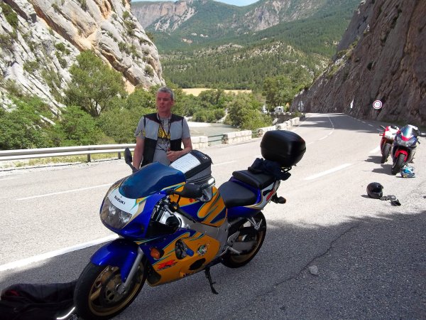 Back from France - Page 1 - Biker Banter - PistonHeads - This image features a man walking alongside a blue, yellow, and orange motorcycle on a mountainous road. The motorcycle is prominently visible in the frame, taking up a significant portion of the image. The man is dressed in casual attire with a vest, necklace, and helmet in hand. There are numerous motorcycles parked on the side of the road, suggesting this might be a popular spot for riders. The road curves sharply to the right, with protective barriers indicating the steepness of the incline. The terrain is rugged and rocky, indicative of a mountainous area. The overall scene suggests an adventure or touring event taking place.