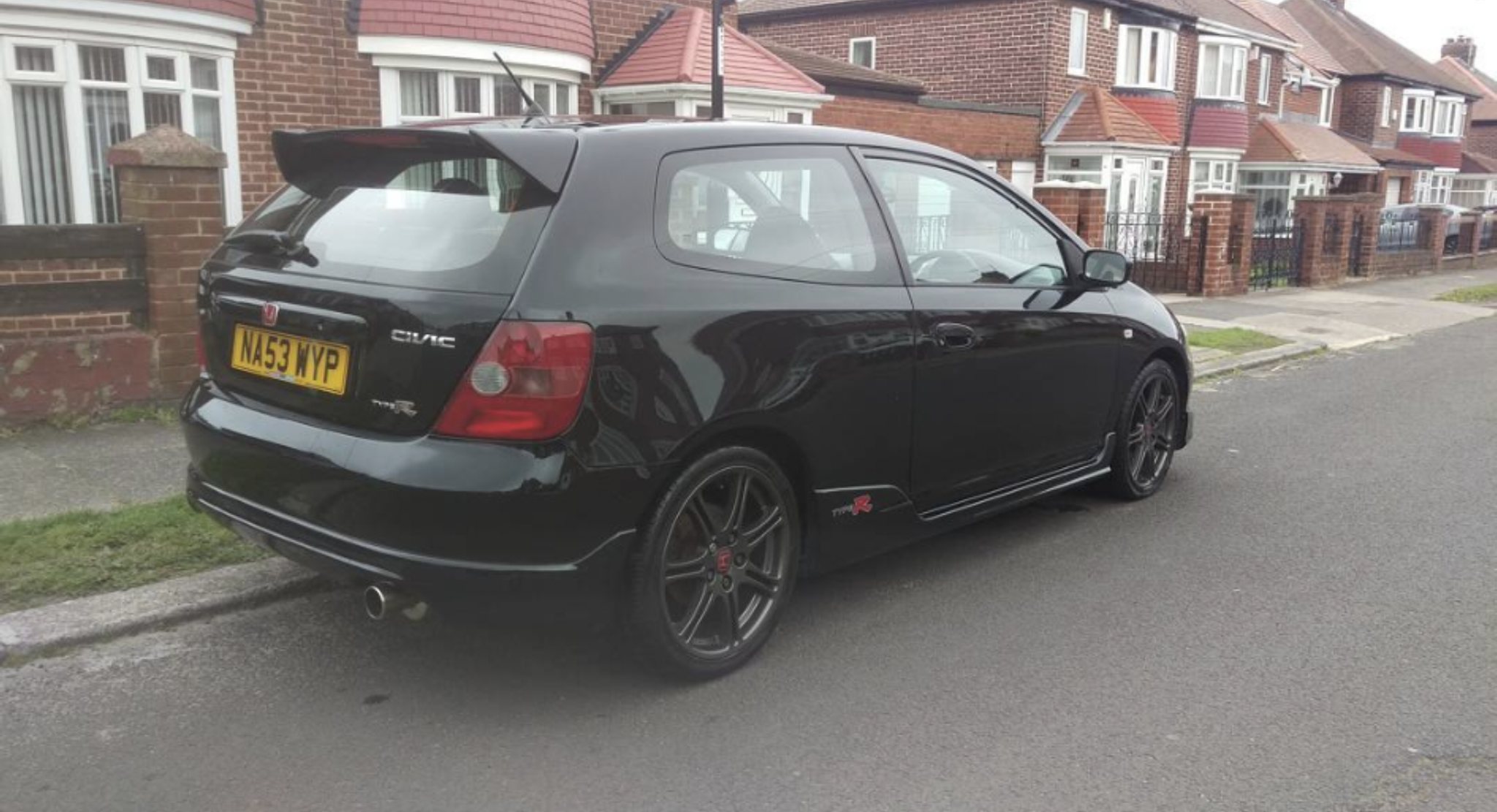 PH Newbie - Page 1 - North East - PistonHeads UK - The image shows a black hatchback car parked on the street. It appears to be a two-door vehicle with a visible spoiler and a rear wing. The car is equipped with aftermarket modifications, such as custom wheels and a performance exhaust system. There are no people in the immediate vicinity of the car. The setting suggests a residential area with houses visible in the background.