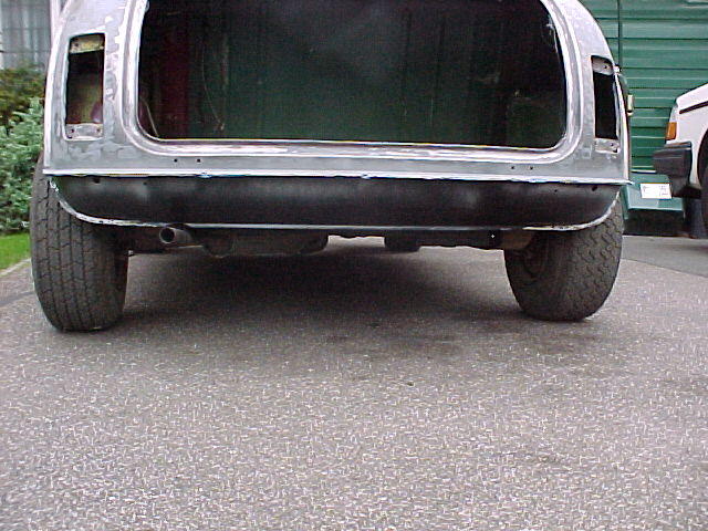 Purchase Pistonheads Advice - The image showcases the rear end of an older model vehicle, specifically focusing on its bumper, which appears to be dented. The bumper is predominantly gray with dark gray elements, possibly indicating some wear or damage. The tires of the vehicle can be seen, with the rear one being more visible. The vehicle is parked on a gray paved surface, with its tail-lights facing us. In the background, there's a hint of greenery, suggesting the location could be near a park or a garden. The overall scene is rather ordinary, capturing a quiet moment of a car waiting in a lot.