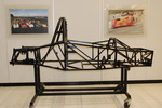 Clubsport Converting Seater Pistonheads - The image features a racing car model mounted on a stand, likely displayed in a gallery setting. The car itself is intricate, with a detailed and realistic design, showcasing its black metal structure that simulates the lines and curves of a racing car. The model is placed on a white tile floor, and there are two pieces of artwork on the gallery walls with visible frames and paintings, indicating an indoor exhibition space. The background is neutral, with walls painted in a light color, providing a contrast that highlights the model.