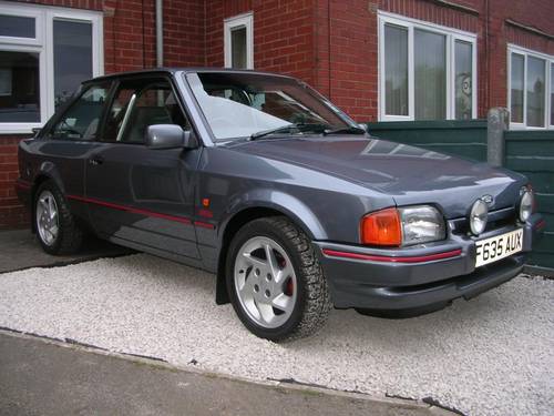 Which car best sums up the 1980s? - Page 7 - General Gassing - PistonHeads - The image features a steel-colored second-generation Volkswagen Golf GTI, identified by a badge on the front car panel. The car is parked on a white pebble driveway in front of a brick wall and a house with multiple windows. It appears to be in good condition, suggesting it is well-maintained.