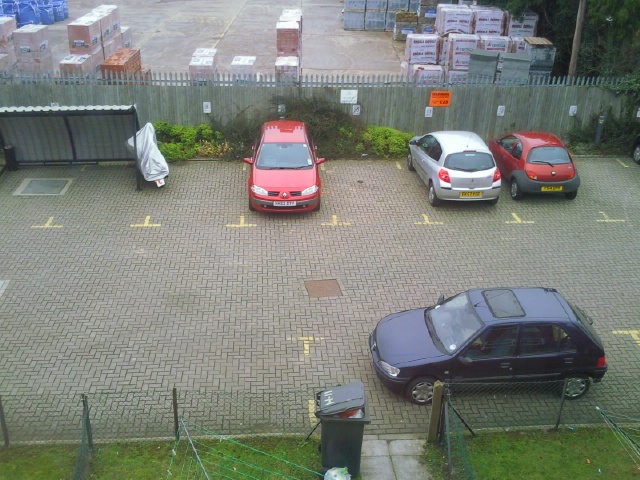 Pistonheads - The image showcases a parking lot with four parked cars, two facing us and two facing away. The lot is paved with red bricks, providing a distinctive backdrop. In the left corner, a brown car is parked next to a black bin, while a blue car is parked in the right corner. Two red cars are parked amidst the other vehicles, creating a vibrant contrast with the black bin and the brick pavement. The parking lot is surrounded by a sturdy metal fence, suggesting it is a secure parking area.