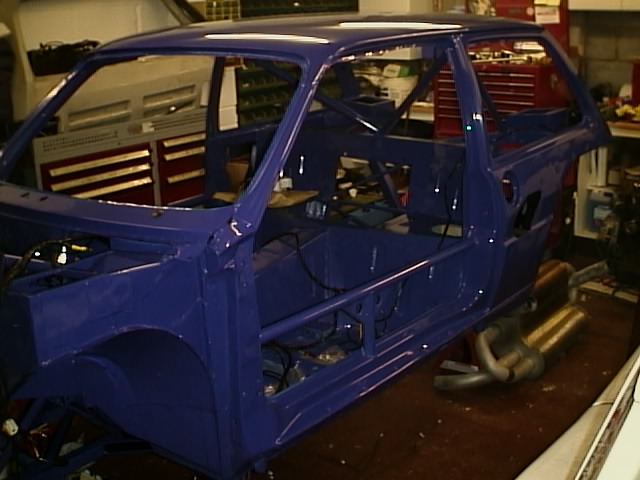 MG Metro 6R4- Under The Plastic - Page 1 - Classic Cars and Yesterday's Heroes - PistonHeads - The image shows a blue car inside a workshop. The car appears to be a hatchback type vehicle, with the rear hatch open revealing its empty interior. The interior is visible through the missing panels and the exposed wires suggest that some work has been done or is in progress. The image has an industrial look due to the focus on the vehicle and the surroundings. There are indicator lights illuminated in the background, which suggests that some electrical work may be occurring.