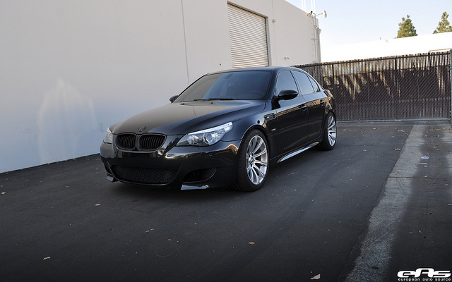 A car parked in front of a parking meter - Pistonheads - The image features a black BMW sedan parked on an asphalt surface next to a parking lot light pole. The car is positioned facing the camera, showcasing its sleek design and distinctive BMW headlights. The parking lot appears empty, emphasizing the car's solitary presence. A warehouse or industrial building forms the backdrop, suggesting an urban or commercial setting.