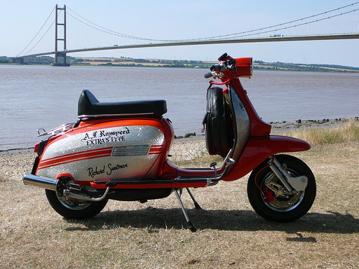 Show us your scooter thread - Page 4 - Biker Banter - PistonHeads - The image shows a vintage red and white scooter parked on grass near the water, with a bridge in the background. It features a design resembling a classic American motorcycle, complete with a saddlebag and chrome details. A small text above it reads "R&H Customs", suggesting it may be a custom or restored scooter. The setting suggests a leisurely, possibly touristy location, given the presence of water and the casual placement of the scooter. The image has an overall nostalgic vibe due to its subject matter and composition.