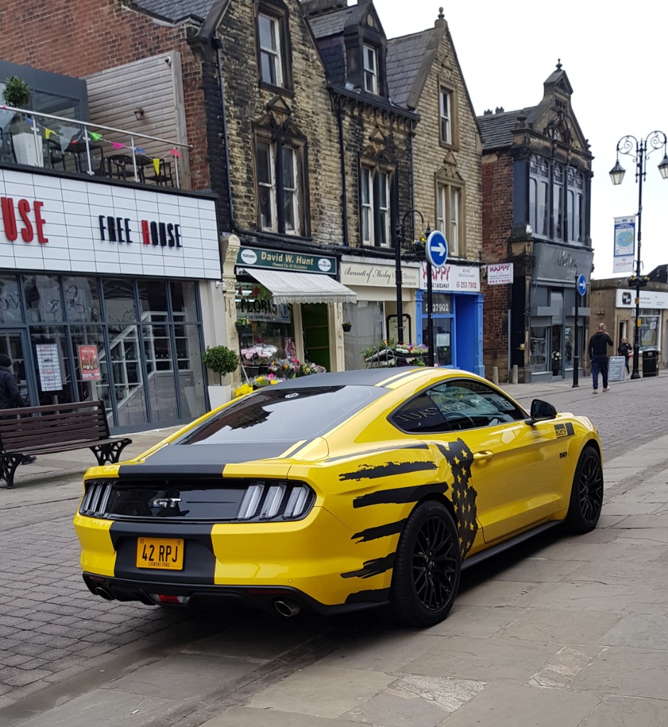 What C124PPY personalised plates have you seen recently? - Page 451 - General Gassing - PistonHeads - The image shows a vibrant yellow Mustang car parked on the side of a street. The vehicle is adorned with black and silver stripes running along its body, adding to its striking appearance. Its design features a prominent hood scoop, which is indicative of high-performance models. In the background, there's a quaint European-style town with buildings and storefronts lining the street. The scene is peaceful, with no people or movement visible in the image.