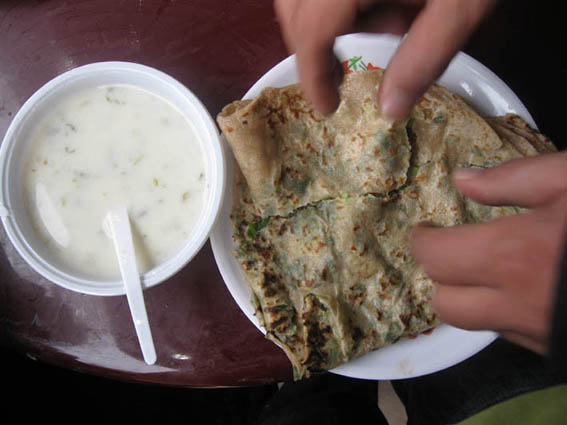 The image depicts a close-up view of a person in the process of enjoying a meal. A hand is reaching out to grab a piece of flatbread, while a bite has been eaten from it already. The flatbread has a golden-brown color and is partially covering a soupy dish. The discarded silverware lies in the starkly contrasting white container adjacent to the main plate. It appears to be a casual dining scene, perhaps someone home cooking.