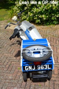 A 'period' classics pictures thread (Mk III) - Page 16 - Classic Cars and Yesterday's Heroes - PistonHeads UK - The image displays a vintage-style motorcycle parked on a brick surface. In the foreground, there is a blue crate or basket attached to the front of the bike. To the right of the motorcycle, there's a text overlay that reads "Lean like a GP rider" in white font on a black background. The motorcycle has a prominent front wheel and a small, round headlight. The background is blurred but suggests an outdoor setting with vegetation.