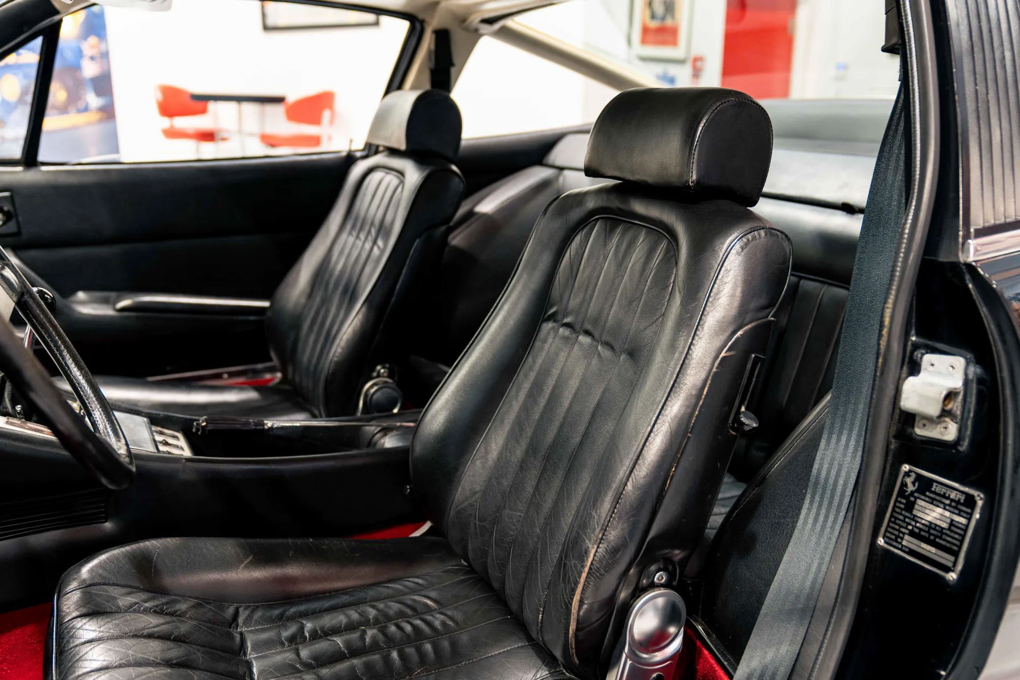 365 GTC/4 - Page 1 - Ferrari Classics - PistonHeads UK - The image captures the interior of a classic muscle car, showcasing its vintage charm. The vehicle is parked inside a building that appears to be a garage or auto showroom. 

The car's black leather seats are the focal point, inviting viewers to imagine themselves in the driver's seat. A red floor rug adds a pop of color to the scene, contrasting with the otherwise monochromatic interior. 

The dashboard and steering wheel are clearly visible, hinting at the car's powerful engine and advanced technology. Despite being an old model, the car maintains its pristine condition, reflecting its well-cared for history. The overall composition of the image exudes a sense of nostalgia and automotive passion.