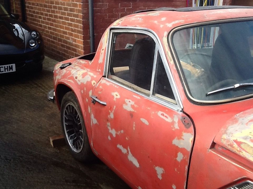 Early TVR Pictures - Page 121 - Classics - PistonHeads - The image captures a scene featuring two classic cars. The main subject is a rusty red classic car, which appears weathered and shows signs of age. This car occupies most of the frame, filling the left side with its vibrant color and general state of disrepair. In the background, partially out of focus, there's a second classic car. Its color is a much darker, almost black, contrasting with the red car. This second car is situated just behind the red one and also presents some noticeable rust spots on its paintwork. The presence of two classic cars in close proximity adds a narrative element to the image, suggesting a shared history or perhaps the remnants of a past collection.