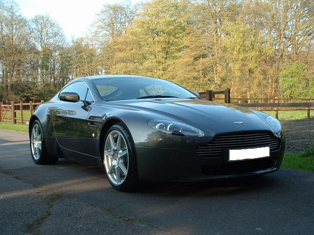 Pistonheads Impressions Months Pros Cons Ownership - This is a shiny, black luxury car parked on a concrete area next to a grassy field filled with trees. The car is sporting a pair of alloy wheels and features a sleek design, typical of high-end vehicles. The Aston Martin logo is prominently displayed in the center of its grille, indicating the brand of the vehicle. The background suggests a residential or rural environment, possibly a driveway or a parking lot.