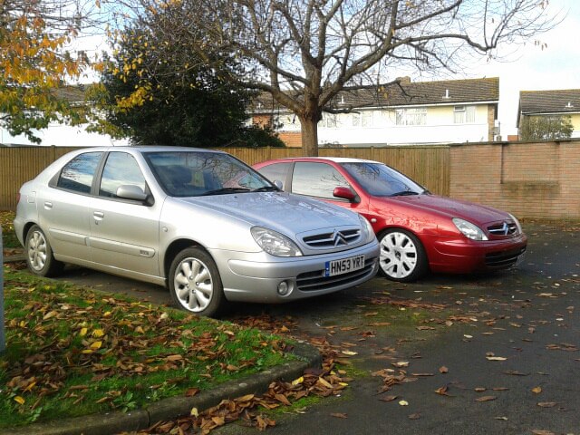 My Two French beauties. - Page 2 - French Bred - PistonHeads - The image shows two cars parked side by side on grass. The car on the left is silver, while the one on the right is red. The cars appear to be parked in a suburban driveway, as there is a wooden fence visible in the background. The surrounding area is grassy with leaves scattered across it, suggesting it might be autumn. No people are visible in the scene, and the focus is on the two parked cars.