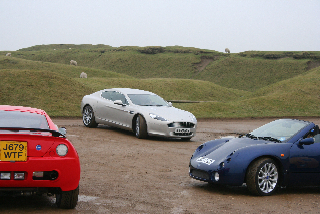 Spotted Aston Rapide Pistonheads - The image depicts a trio of luxury sports cars parked on a dirt path. The backdrop features a pastoral scene with rolling hills blanketed in green grass, providing a stark contrast to the sleek, high-performance vehicles. The road leading to the cars appears to be muddy, suggesting recent or upcoming rainfall. The design and finish of the cars imply that they are luxurious models from prestigious manufacturers. The overall ambiance of the image is serene, with a sense of solitude and tranquility.