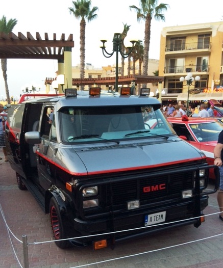 In Malta last week - Page 1 - Yank Motors - PistonHeads - This image captures an outdoor scene where a black GMC truck is parked on a tiled area next to a red sports car. The truck is lifted and features a modified design with extensive visual modifications. Its grille is customized and has the word "GI MOB" prominently displayed, suggesting that the truck may be associated with a custom automotive company or a group that specializes in vehicle enhancements. In the background, there is a pedestrian area with palm trees, and several people are observing the modified vehicles, adding to the atmosphere of this event.