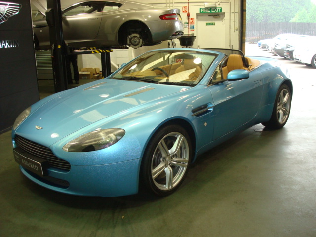 Drove a V8V Roadster - Page 1 - Aston Martin - PistonHeads - The image is a photograph of a sports car in a garage setting. The car is predominantly blue with silver accents, reminiscent of a convertible model. It is positioned on a two-post car lift, which is retracted to the floor as the car is not in use. The surrounding environment is industrial, with what appears to be a workshop door in the background. The floor shows reflections from the environment, suggesting a well-lit and possibly polished area. There are no visible texts, logos, or distinctive brands that provide additional information about the location or the car itself.