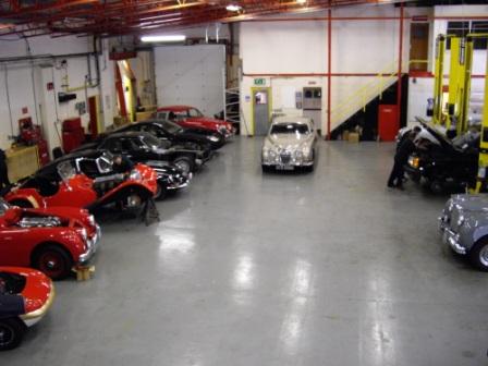 Pistonheads Lotus Workshop Jaguar Projects - The image showcases a spacious garage, amply illuminated with natural light. The floor, made of concrete, is currently free from vehicles, providing a clear view of the surrounding area. At the center of the room is a set of stairs, quietly observing the scene from its elevated position. Scattered across the garage floor are several cars, each part of a different collection. Some cars are facing each other, engaging in a silent conversation, while others stand facing the same direction.