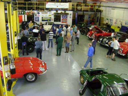 Pictures from Paul Roach's Open Day - Page 1 - Classic Cars and Yesterday's Heroes - PistonHeads - The image portrays a bustling scene inside a large warehouse-like building. A diverse array of old-time cars is parked along the walls and in the center, drawing a crowd of people. The individuals, some clustered in groups, are seen closely examining the vehicles, their attention focused on the rich details of the vintage transportation. The vehicles themselves range from sleek red sports cars to green convertibles, all seemingly in pristine condition. A red car is being lifted by a yellow hoist, hinting at some maintenance or installation work underway in the spacious garage. The atmosphere of the scene suggests a car show or exhibition, with the vintage cars as the main attraction.