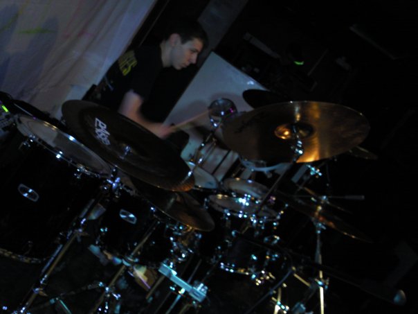 Pistonheads Lets Gig - In the image, a man is actively playing a drum set. He stands at the center, immersed in his performance. The drum set is well-equipped and positioned in the center of the room. There's a cymbal and a large black drum nearby, as well as other silver cymbals to complete the setup. The scene suggests a lively performance at a dark studio or venue.