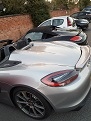 Boxster Spyder - Page 432 - Boxster/Cayman - PistonHeads - The image shows a row of parked cars on a street. The cars are side-by-side in what appears to be a parallel parking row, with various models and colors. The car in the foreground, which is silver and red, is a sports car with a sleek, sporty design, featuring a low roofline and a spoiler. The car's lighting and bodywork appear polished, indicative of possibly being a convertible or a high-end sedan. The street is lined by a brick wall to the left, suggesting an urban setting.