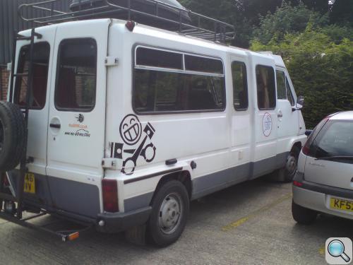 Pistonheads - This image features a white van parked in a lot. The van appears to be modified, with a large rack mounted on top and a small platform at the front. On the side of the van, there are decorative graphics and text, along with some smaller decals that seem to feature stylized drawings, possibly related to the vehicle's function or the company it represents. The van is closely parked next to a smaller car, suggesting a shared space or parking requirements specific to the site. The surroundings show other vehicles and vegetation, indicating an outdoor parking lot.