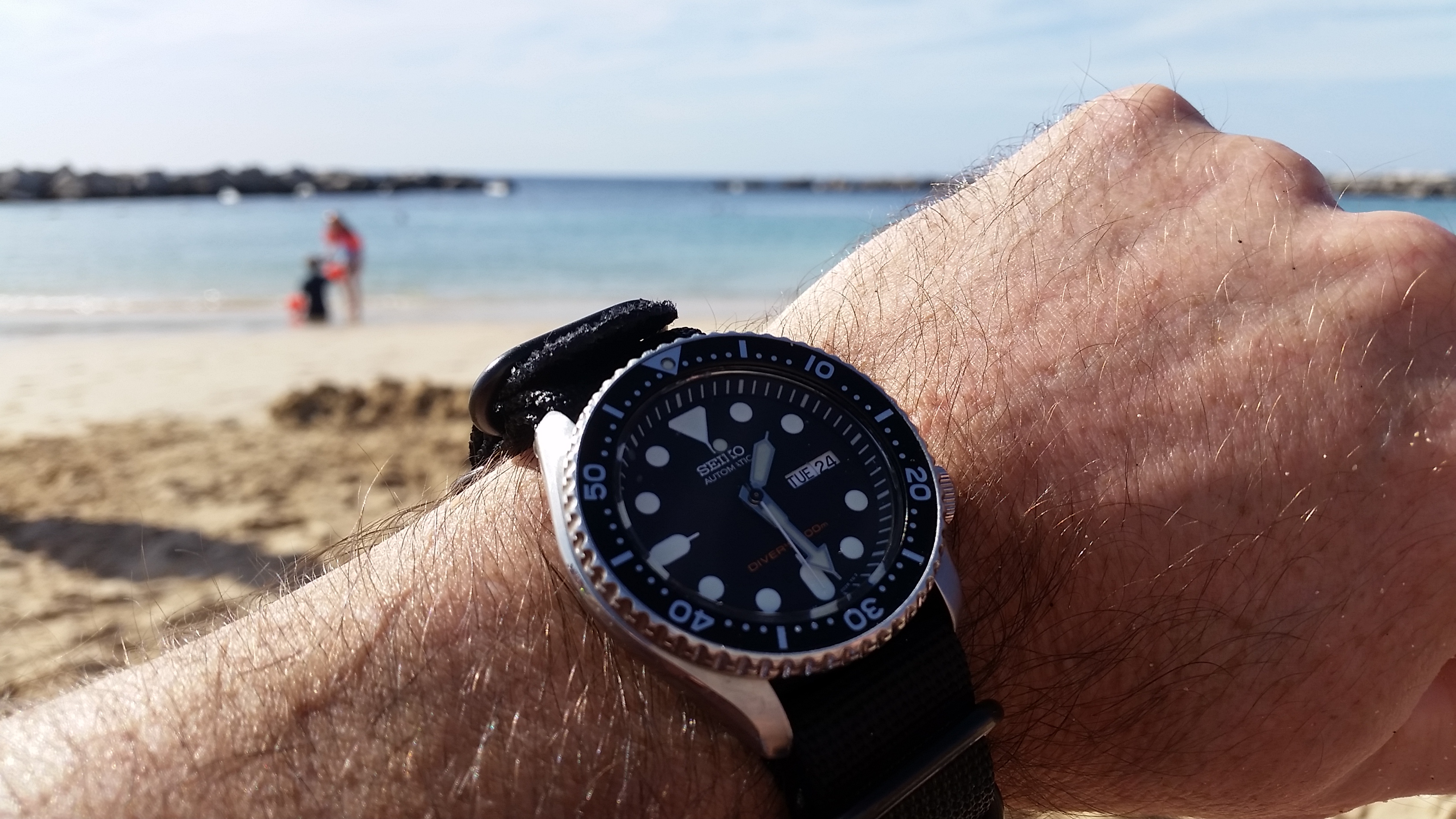 Let's see your Seikos! - Page 133 - Watches - PistonHeads - The image shows a person's arm holding a wristwatch. The watch appears to be black and white with a strap, likely made of leather or a similar material. The individual is wearing a band around the watch face, which might be used for measuring heart rate or other fitness metrics. In the background, there is a beach scene with clear blue skies and calm waters in the distance. There are also some rocks visible near the water's edge.