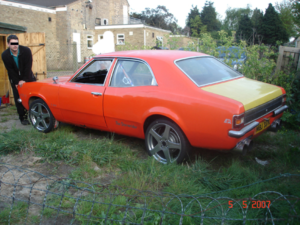 Orange Cars - Page 7 - General Gassing - PistonHeads - In the image, a man is standing next to an orange car that appears to be a classic from the 1960s or 1970s. The car is parked in a grassy area near a fence. The man is viewing the coupe, which has a vibrant color, a prominent body kit, and a yellow rear section with a black stripe in between. The setting seems to be a suburban area with houses in the background, suggesting a residential neighborhood.