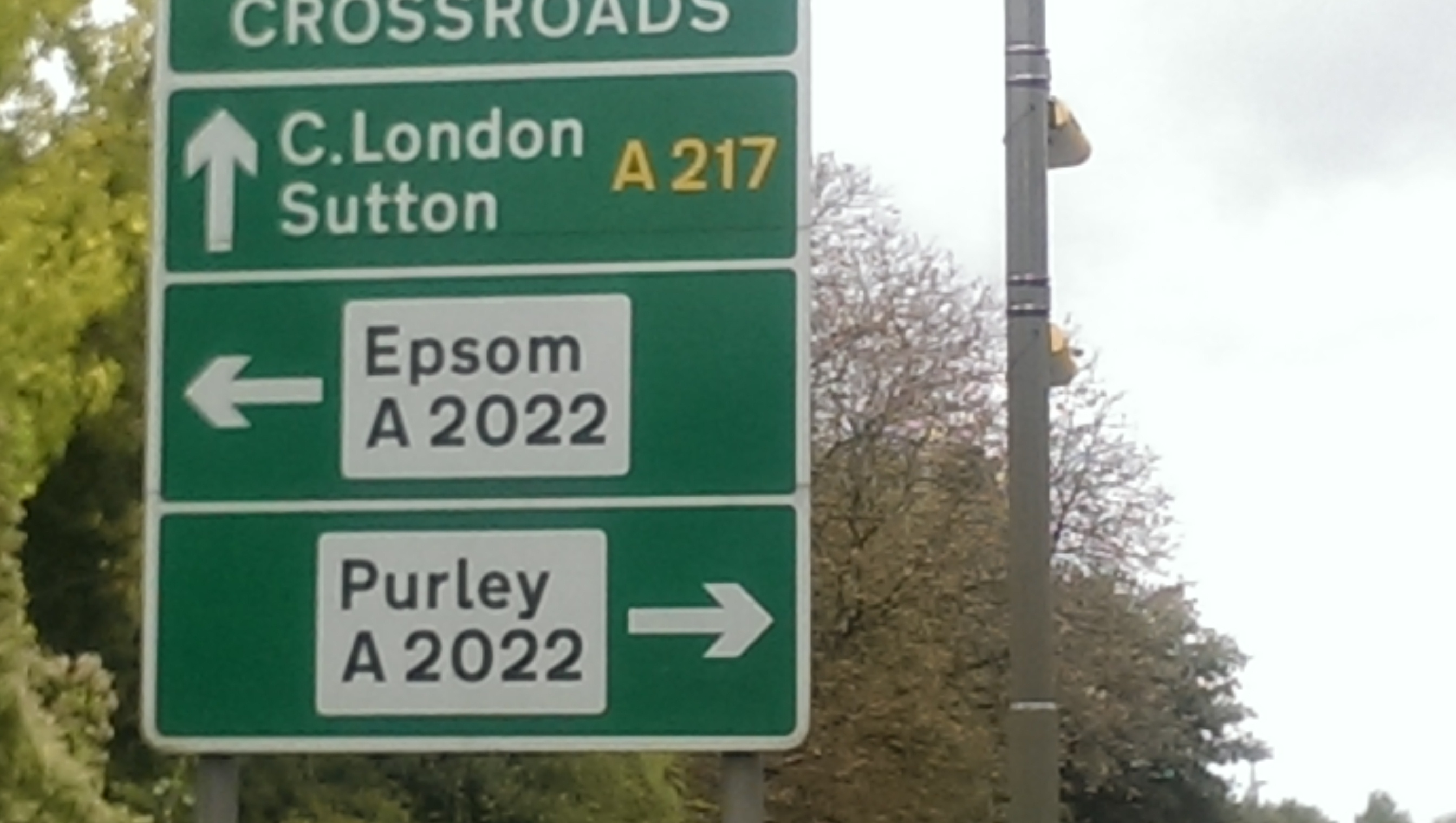 WARNING: A217 Reigate to Sutton  New Average Speed Scameras - Page 13 - Roads - PistonHeads - The image shows a green street sign by the roadside. The sign displays directions with white text and yellow numbers. The top sign points toward "C.London Sutton A217" and the bottom sign indicates "Espom A2022 Purley A2022." There are trees and a clear sky in the background, suggesting a day with good weather. The signs provide guidance to drivers for the correct route to take.