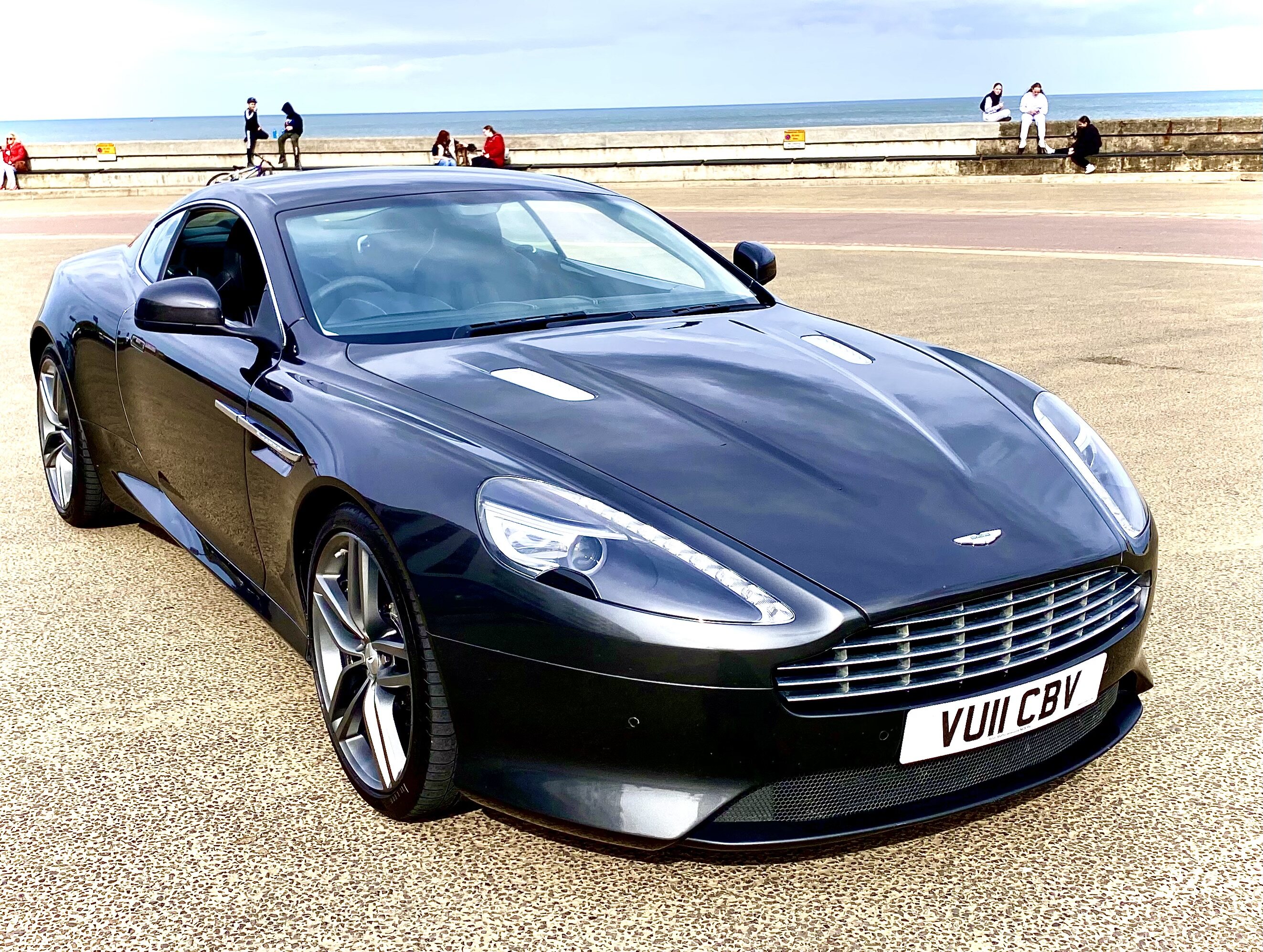 Pistonheads - The image depicts a black sports car parked on what appears to be a beachfront road. The car is a convertible with the top down, showcasing its sleek design and shiny exterior. It has prominent side vents that are characteristic of high-performance vehicles. There is visible branding on the hood and wheels, indicating it's an Aston Martin model. In the background, there are people and buildings, suggesting a coastal town or city area. The overall scene is bright and sunny, with clear visibility of the car and its immediate surroundings.