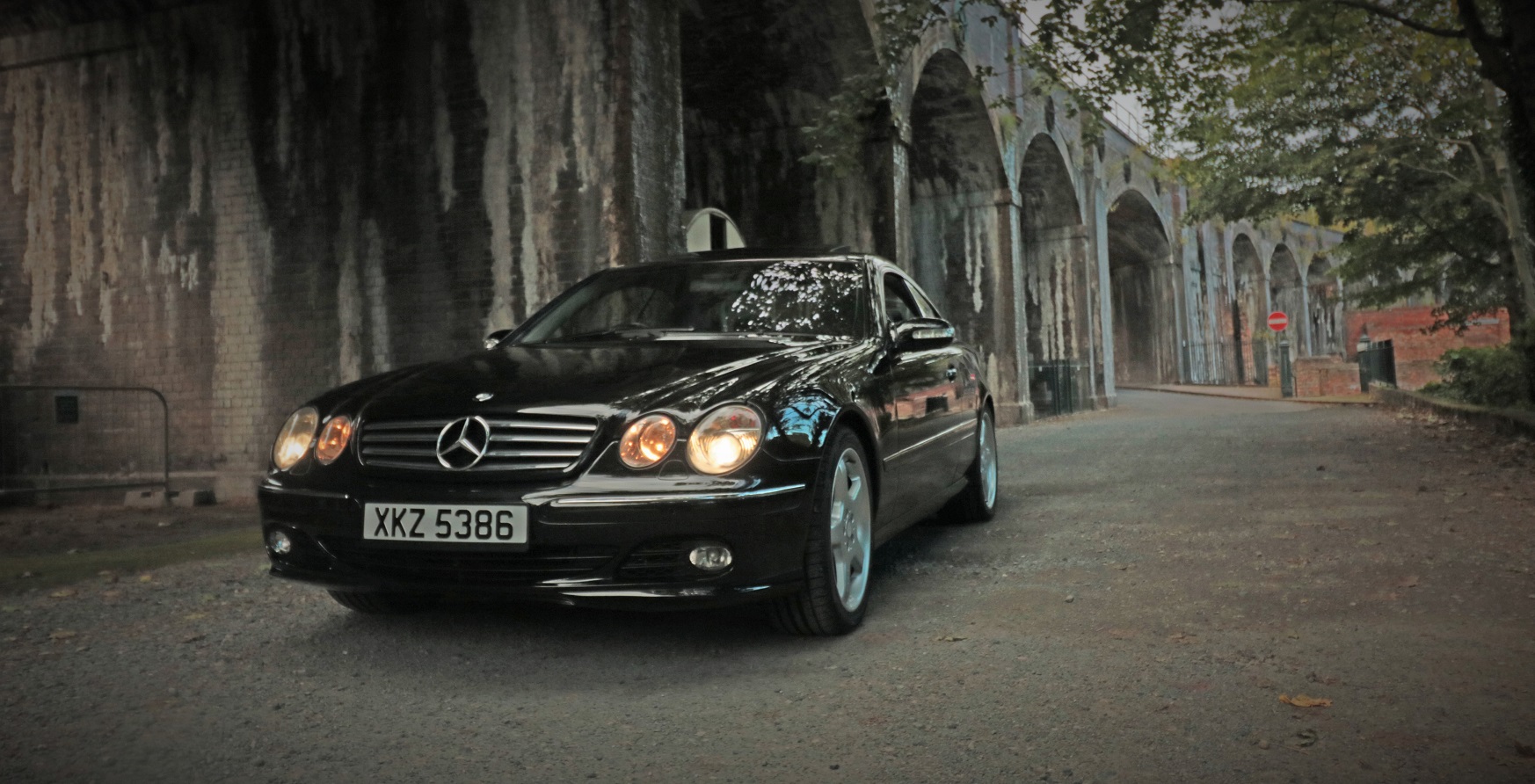 The Tank - CL500 - Page 2 - Readers' Cars - PistonHeads - The image is a photography featuring a black Mercedes-Benz car parked on a street during twilight or what appears to be dusk. The car is positioned to the left of the frame, portraying its side profile. The car's headlights are illuminated, and through them, the reflection of trees can be seen. Overhead, the structure of an arched bridge or walkway is visible, suggesting that the road may be an overpass or that this is a photo taken under a bridge. The surrounding environment is dimly lit, and there is cobblestonework visible, adding to the scenic and somewhat moody atmosphere of the image.