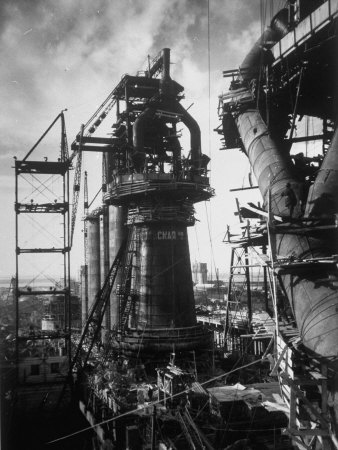 Objects Evil Houses Eerie Inanimate Pistonheads - This black and white photo captures a scene at a steel mill. The mill, towering and imposing, dominates the scene with its large framework and the silhouette of its tall ladder seen from below. The mill is bustling with activity, hinted at by the figure of a worker visible in the frame. In the foreground, smaller structures add depth to the image, with overhead cranes, and buckets ready for use in the industrial operations. The image transports us to an era of heavy industry, a testament to human ingenuity and the power of technology.