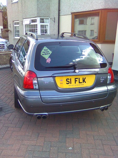 The joys of a big V8 - Page 2 - General Gassing - PistonHeads - In the image, a car is parked on the side of a brick-paved street. The car is a hatchback sedan, painted in a metallic shade that reflects the outdoor lighting. The car's registration plate is clearly visible and reads "SI FLIK." The hatchback appears to be parked directly in front of a house or residential building. The street is relatively quiet with another car visible in the distance.