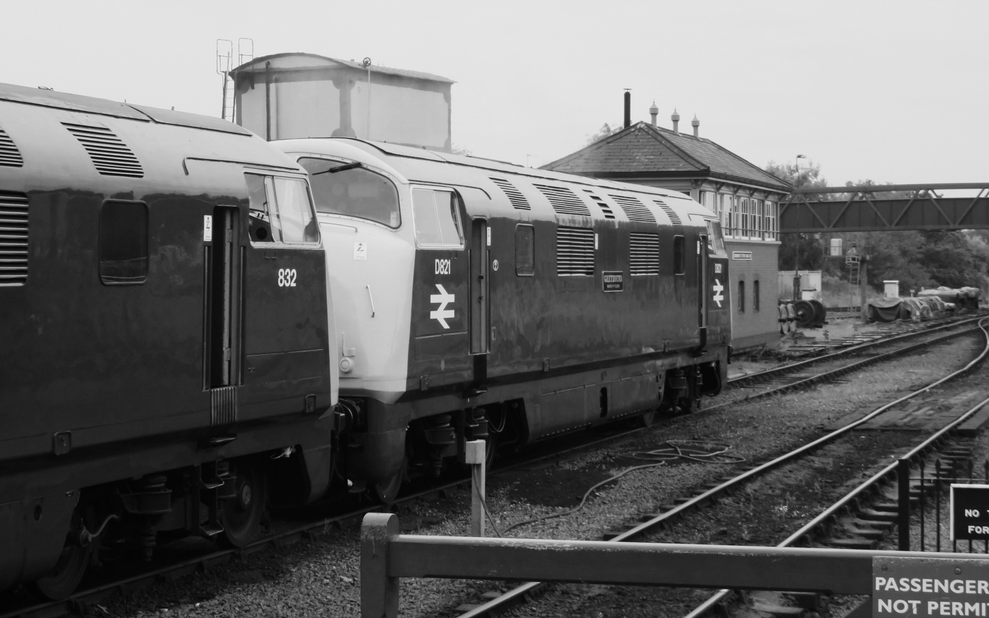 Loco sheds and other railway buildings... - Page 37 - Boats, Planes & Trains - PistonHeads UK - The image is a black and white photograph capturing two diesel locomotives on a railway track. The trains, painted in a shade of green, are stationary with their doors closed. The one on the left is partially visible behind a metal gate, while the other is more clearly seen in its entirety on the right. In the background, there's a building and some greenery, suggesting an urban or suburban setting. There are no people visible in this image, giving it a sense of stillness. The photograph has a vintage feel, possibly indicating it was taken some time ago.