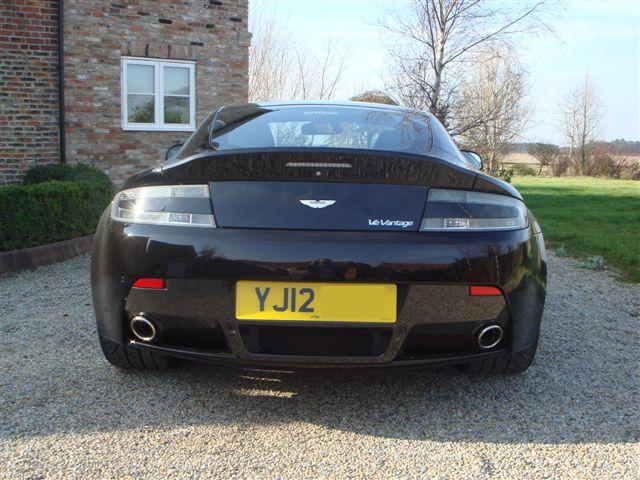 New Vantage V12 - Page 1 - Aston Martin - PistonHeads - The image shows a black Aston Martin car parked on a gravel driveway. The luxurious vehicle is the central focus of the picture, with its glossy finish reflecting the light. The license plate reads "YJ12" and there are no other visible vehicles or people. In the background, a brick house suggests a residential setting, and there are some trees and open fields visible, indicating a peaceful rural area.